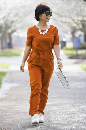 Woman wearing rust-colored cotton linen set with short-sleeve top and straight-leg pants outdoors walking with handbag and sunglasses