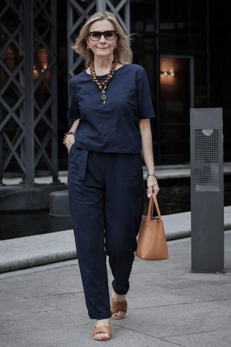 Woman wearing navy cotton linen set with short sleeves and straight-leg pants walking outdoors carrying tan handbag