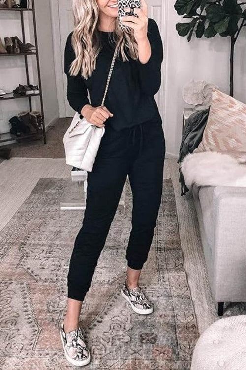 Woman wearing black Cozy Chic Two-Piece Sweatsuit set with long sleeves and pants, paired with snake print sneakers and a white handbag indoors
