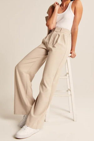 Woman wearing beige Classically Vogue Business Trousers with a white tank top and white sneakers, sitting on a stool.