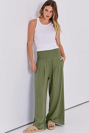 Woman wearing oversized palazzo pants in olive green with white sleeveless top standing against white wall