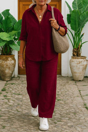 Woman wearing a relaxed shirt and trousers two-piece set in burgundy with white sneakers and a woven shoulder bag outdoors.