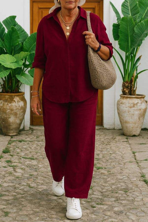 Woman wearing a relaxed shirt and trousers two-piece set in burgundy with white sneakers and a woven shoulder bag outdoors.