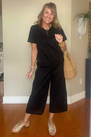 Woman wearing black loose fit co-ord set with short sleeves and wide-leg pants, styled with sandals and a woven handbag.