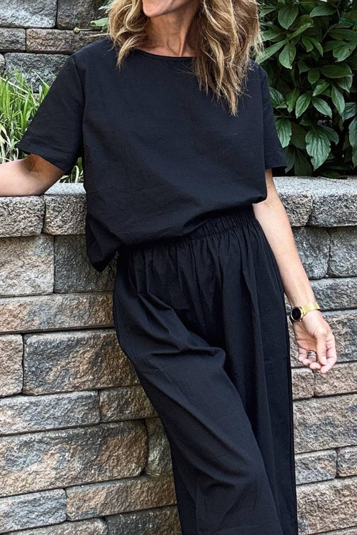 Woman wearing a black loose fit co-ord set with wide-leg pants and a relaxed short-sleeve top against a stone wall background
