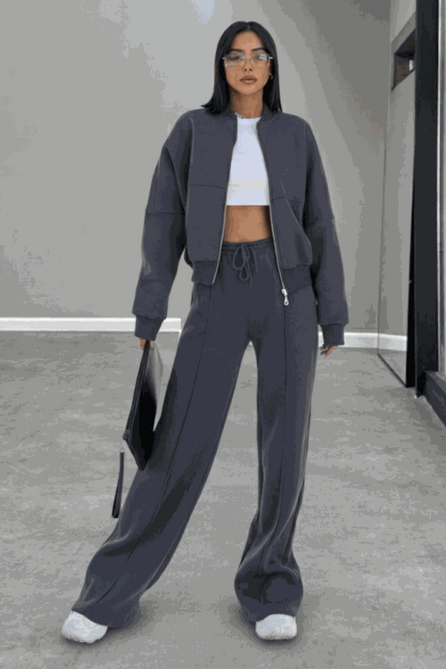 Woman wearing The Voyager Zip Set in charcoal gray, featuring a relaxed zip-front sweatshirt and wide-leg joggers with pockets, styled with white sneakers and a tote bag.