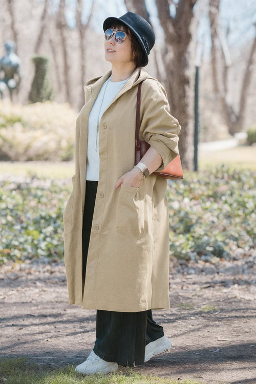 Woman wearing oversized light brown waterproof windbreaker jacket with hood, sunglasses, black hat, white shoes, and carrying a shoulder bag outdoors in a park