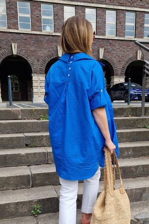 Woman wearing a bright blue oversized linen shirt with back button details walking up outdoor stone steps carrying a woven bag