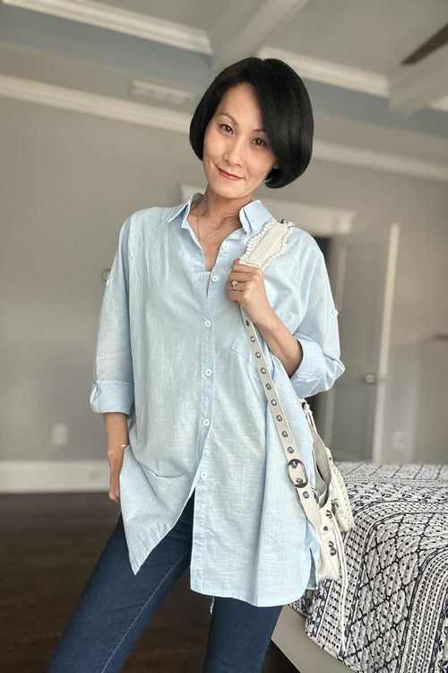 Woman wearing oversized light blue linen shirt with rolled sleeves and white shoulder bag indoors