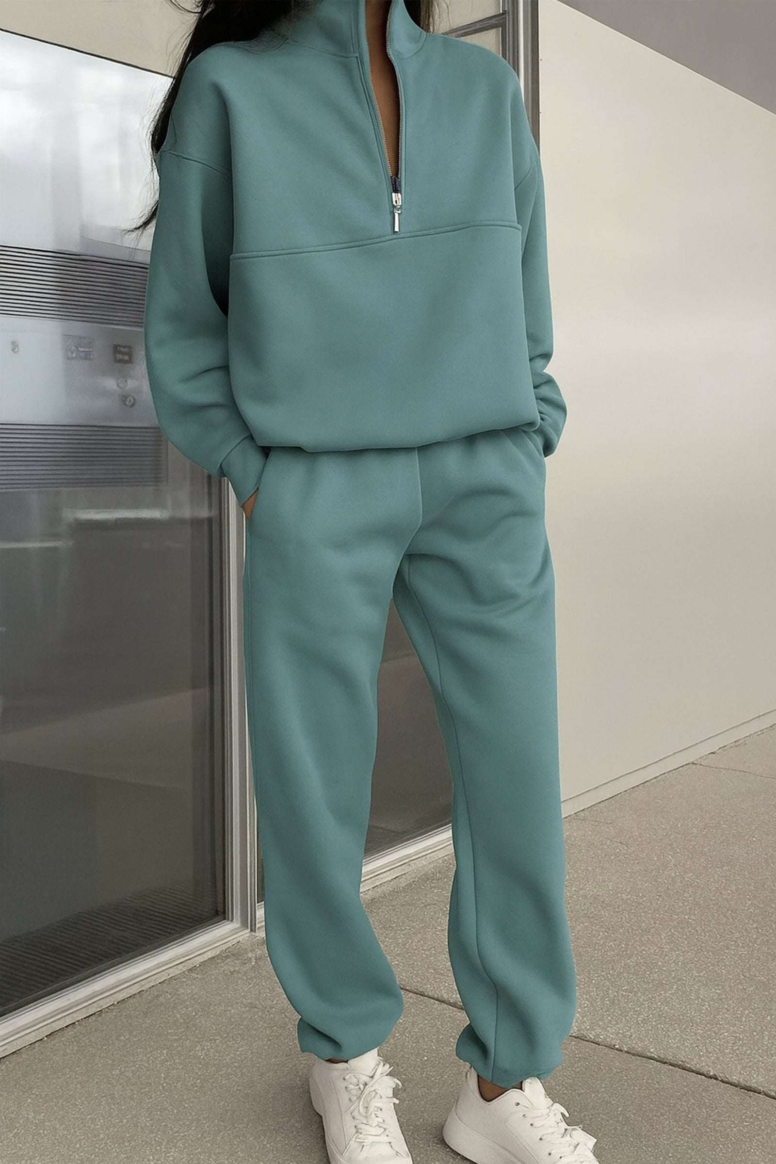 Woman wearing mint green cotton blend tracksuit with zip neckline and white sneakers, standing indoors.