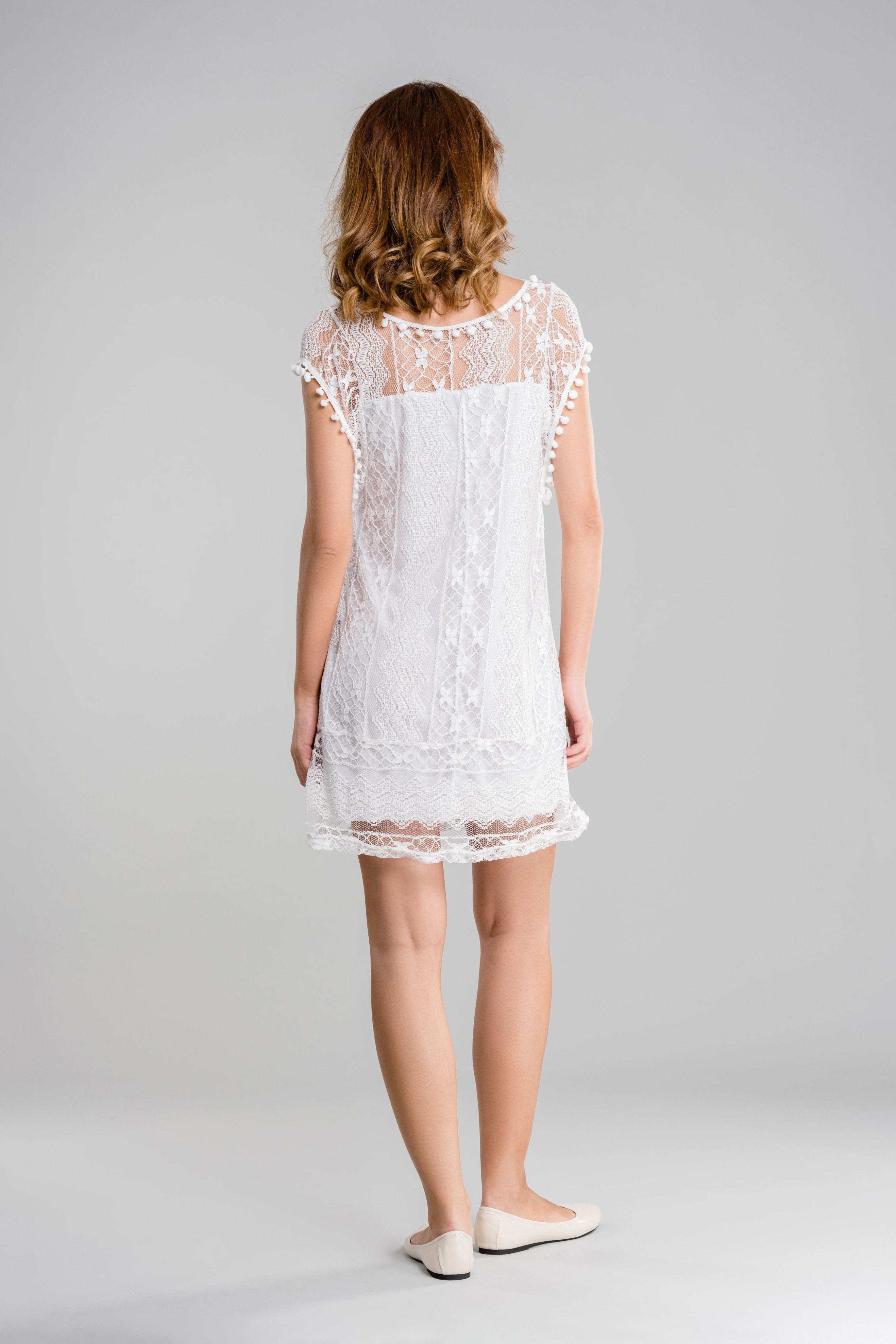 Woman wearing white Pom Pom Lace Mini Dress with sheer lace back and pom pom trim, standing against gray background