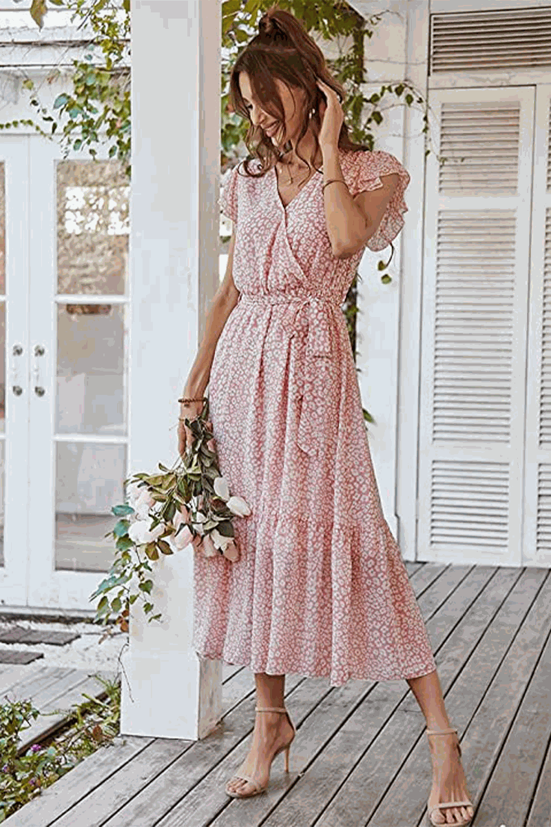 Woman wearing Elegant Blossoms Boho Maxi Dress with floral pattern, V-neck, belted waist, and ruffled hem holding flowers on a wooden porch.