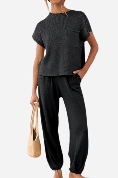 Woman wearing black Comfy Couture Knit Sweater two-piece set with short-sleeve pullover and high-waisted pants, holding beige handbag.
