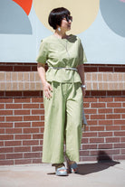Woman wearing light green loose fit co-ord set with a belted top and wide-leg pants standing outdoors against a brick wall and colorful mural background