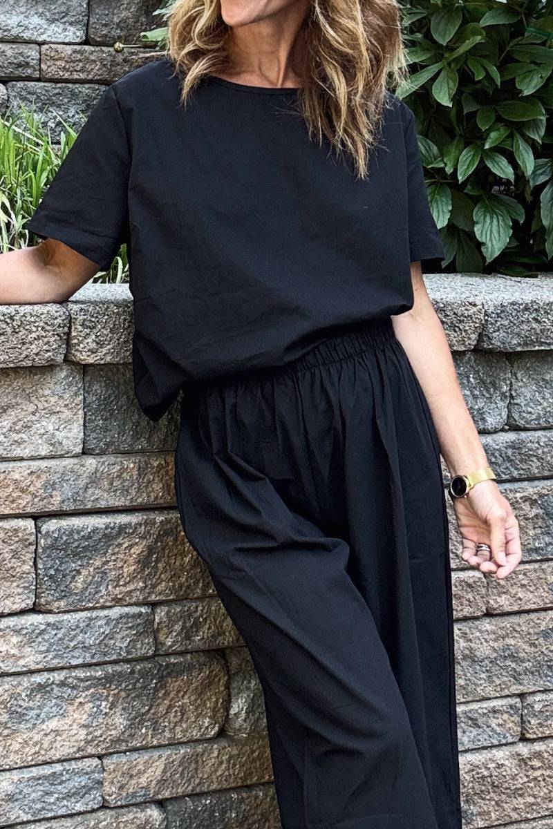 Woman wearing black loose fit co-ord set with short-sleeve top and wide-leg elastic waist trousers outdoors against stone wall