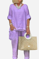 Woman wearing lavender asymmetrical two-piece trouser set with rolled-up sleeves and beige handbag