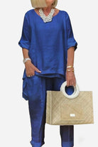Blue asymmetrical two-piece trouser set with rolled-up sleeves and tapered pants, styled with a white beaded necklace and beige handbag.