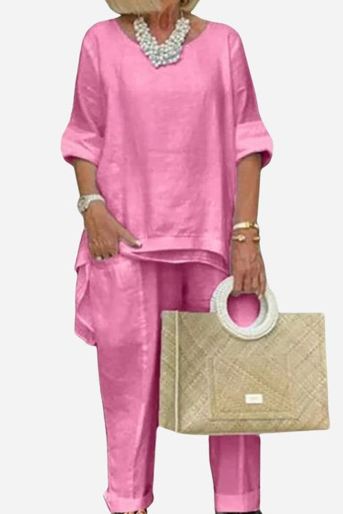 Pink asymmetrical two-piece trouser set with loose top and tapered pants styled with beige handbag and pearl necklace