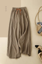 Wide-leg cotton linen Tuscany trousers with elastic waistband and drawstring detail, styled with straw bag and black-tipped shoes on beige background