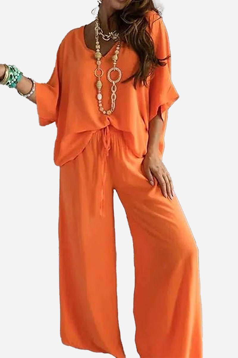 Woman wearing an elegant orange two-piece pantsuit with a V-neck top and wide-leg pants, accessorized with layered necklaces and bracelets.