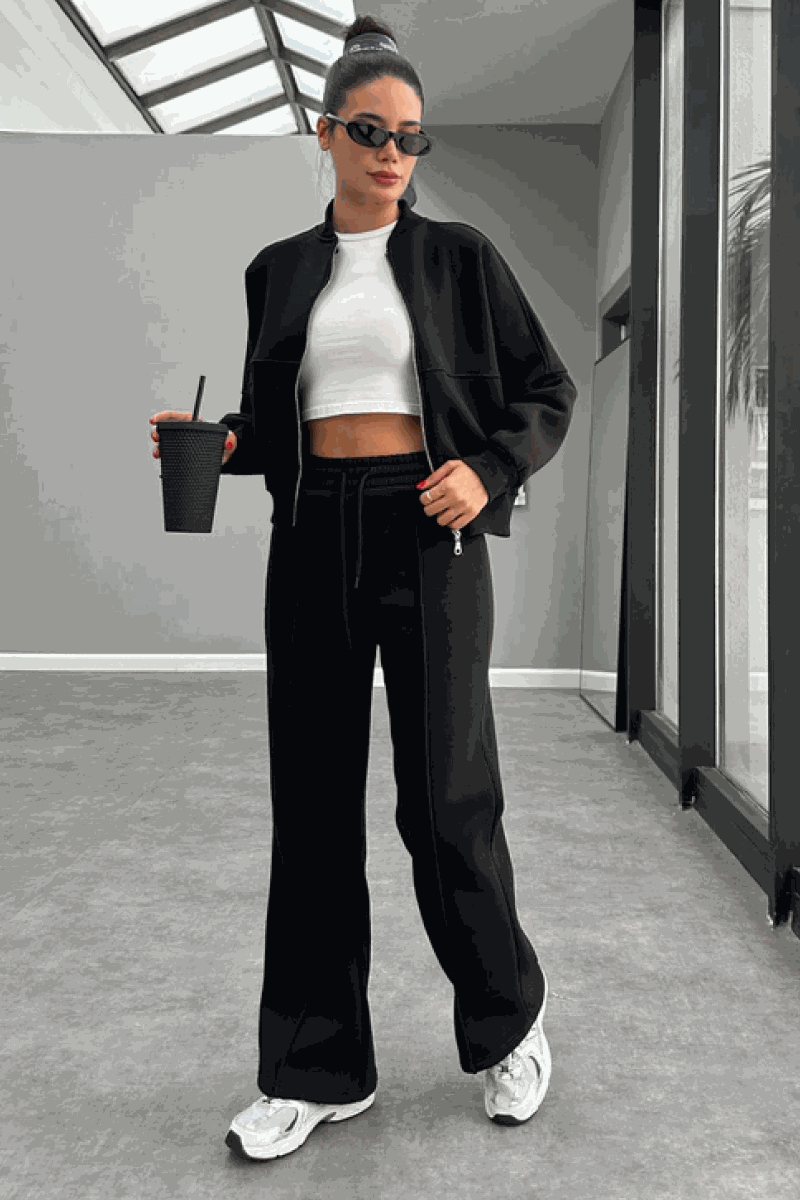 Woman wearing The Voyager Zip Set black zip-front sweatshirt and wide-leg joggers with white sneakers and sunglasses indoors.