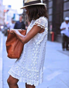 Woman wearing white pom pom lace mini dress with sleeveless design and carrying a brown bag on city street