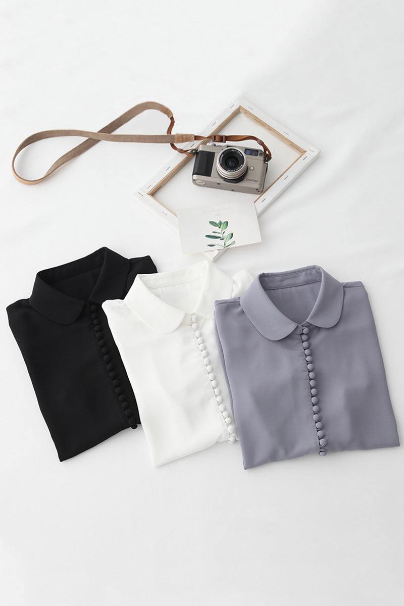 Three folded Bayam Blouse in black, white, and gray with button details and Peter Pan collars displayed on a white surface with a vintage camera and photo frame.