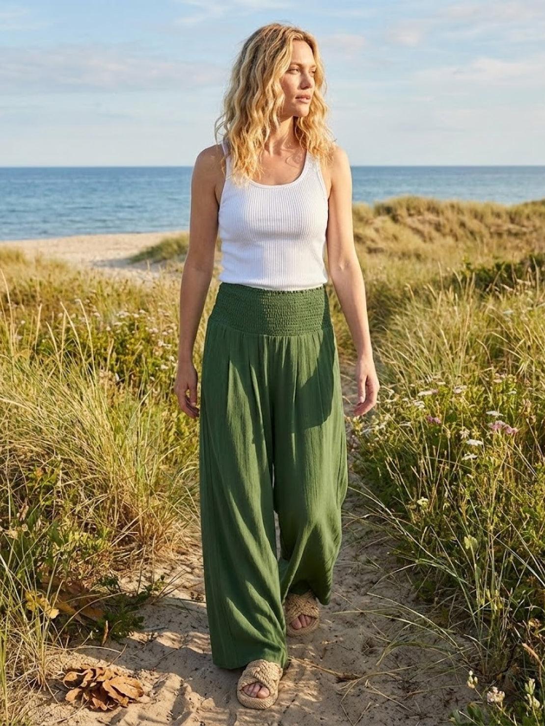 Woman wearing green high-waist palazzo pants and white tank top walking on beach path with grass and ocean background