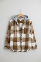 Highland Plaid Shacket with brown and white plaid pattern, soft fleece lining, attached gray hood, button-front, and chest pocket hanging on wooden hanger.