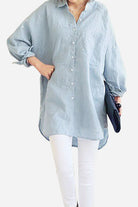 Woman wearing a light blue oversized linen shirt with rolled-up sleeves and white pants, holding a black clutch.
