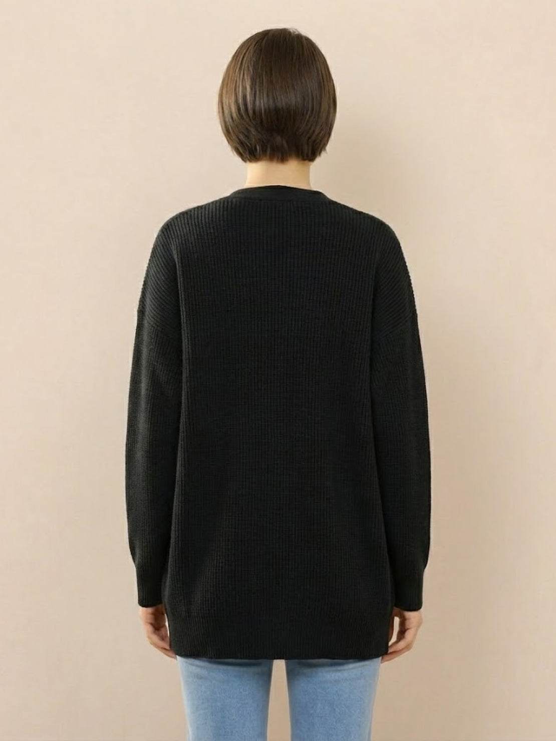 Back view of woman wearing black relaxed fit oversized button down knitwear cardigan with long sleeves and round neckline paired with light blue jeans against beige background