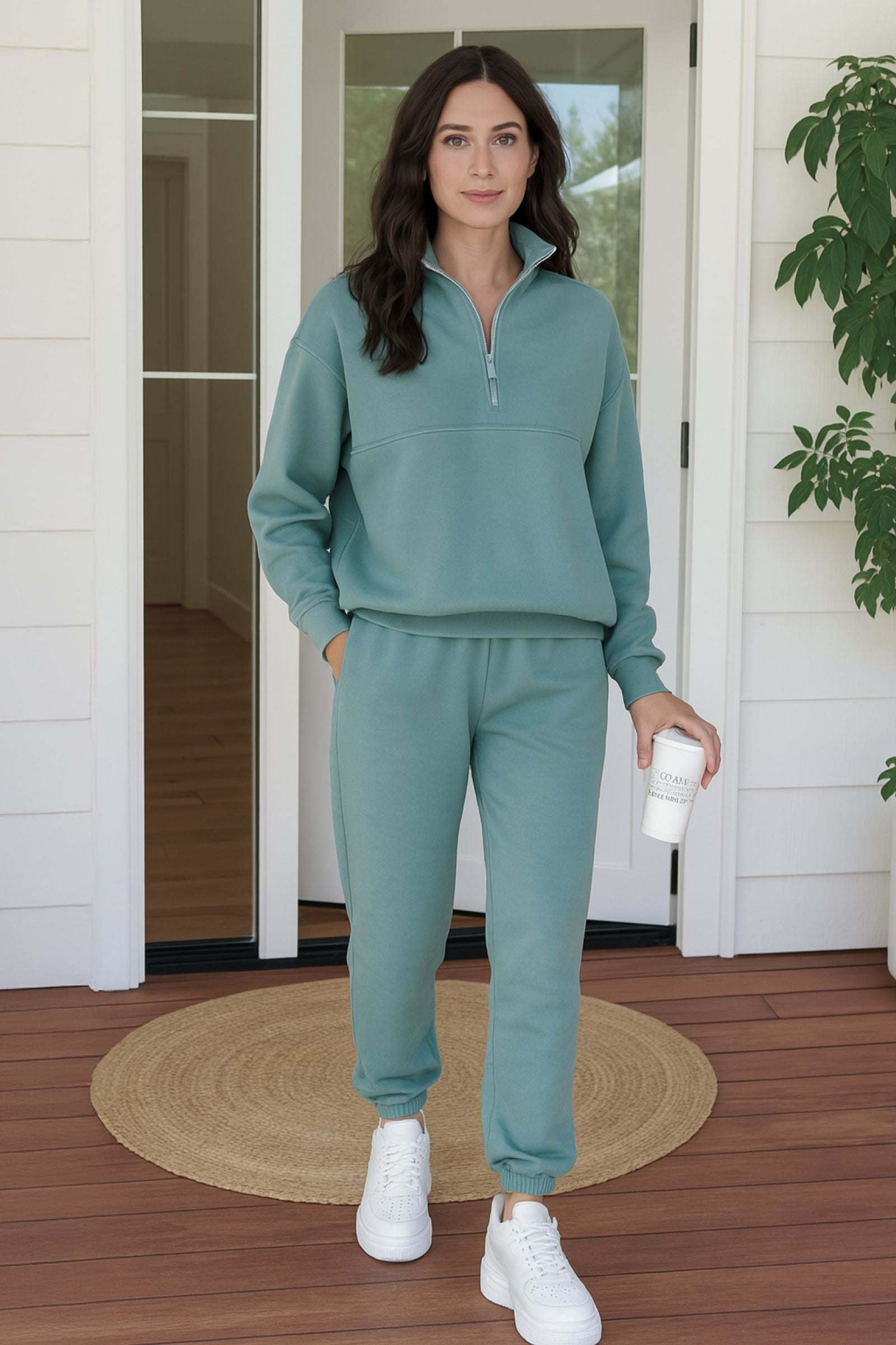 Woman wearing mint green cotton blend tracksuit with zip collar and white sneakers standing on porch holding coffee cup