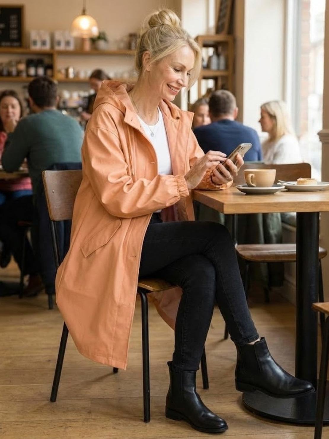 Woman wearing oversized peach Rain Couture Windbreaker sitting in cafe with coffee and smartphone