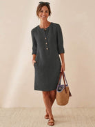 Woman wearing a knee-length dark cotton dress with rolled ¾ sleeves and button front, holding a woven bag.